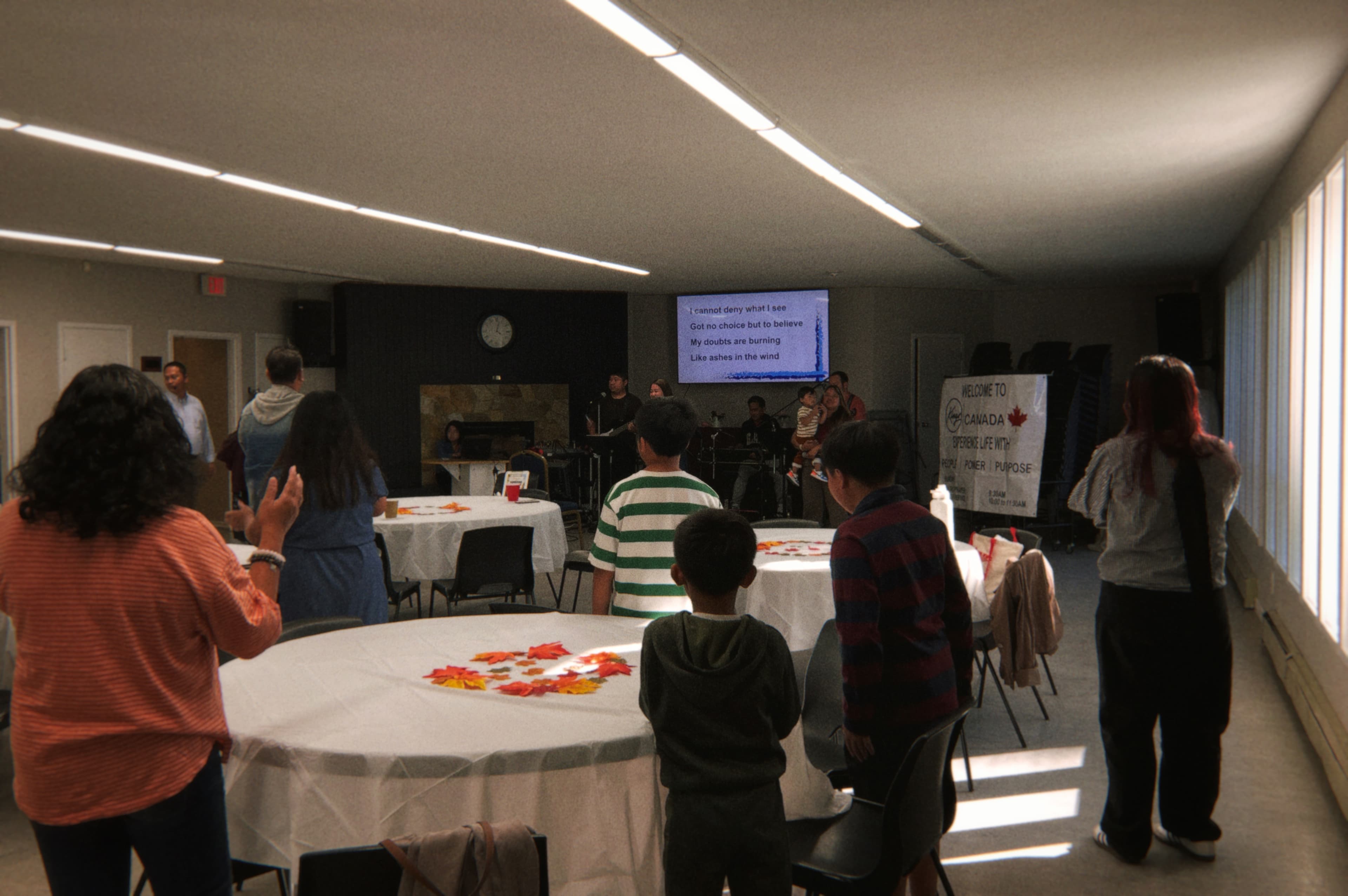 Adults and children standing in a gathering room, facing singers and musicians